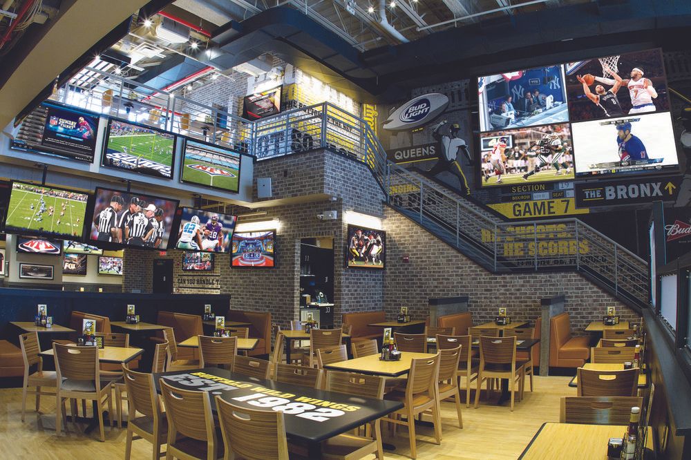 The interior of a two-story sports bar with light brick interior. The walls are covered with posters and television screens.