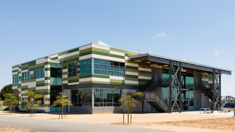 Mount Eden High School, a two-story building clad in Nichiha&rsquo;s Illumination panels in white, light green and dark green is standing in front of a cement walkway and a few small trees.