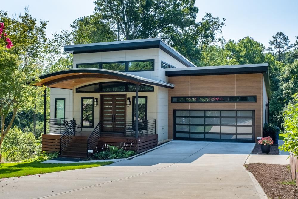 This modern house has an attached garage with wood-like paneling. The front porch has black chairs on it.