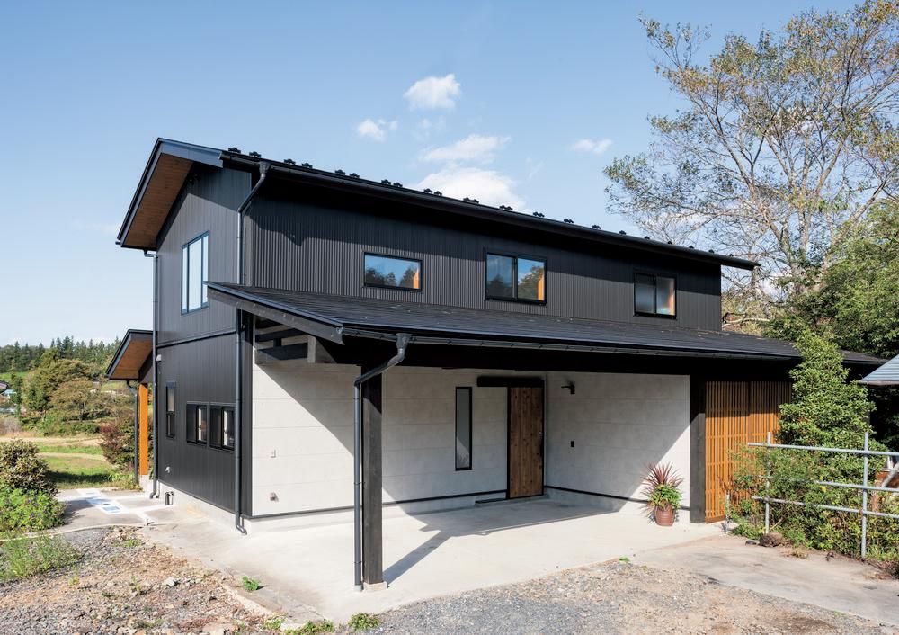 A black and gold wooden home with Nichiha fiber cement siding is the focal point of this photo. There&rsquo;s a blue sky, greenery surrounding and behind the home, and a carport with a white interior.