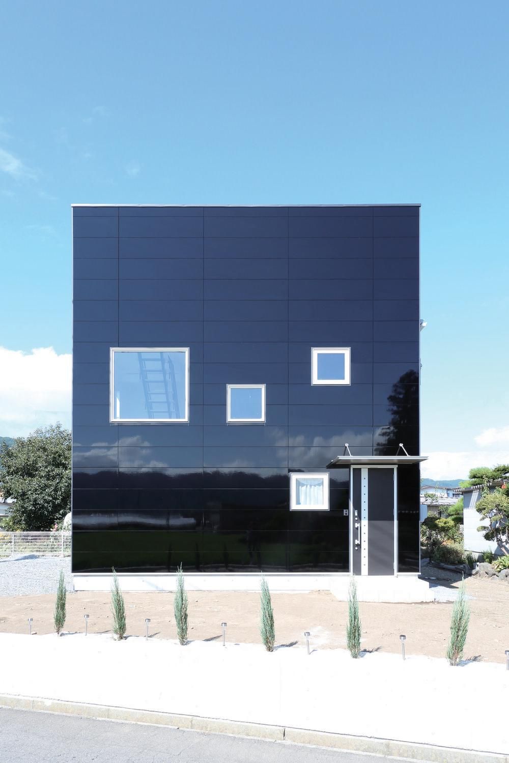 A glossy, black home in Nichiha Miraia siding is the focal point of this image. The house has a door on the right and four differently-sized square windows. There&rsquo;s a blue sky and a few thin, tiny bush trees in front of the house by the sidewalk.