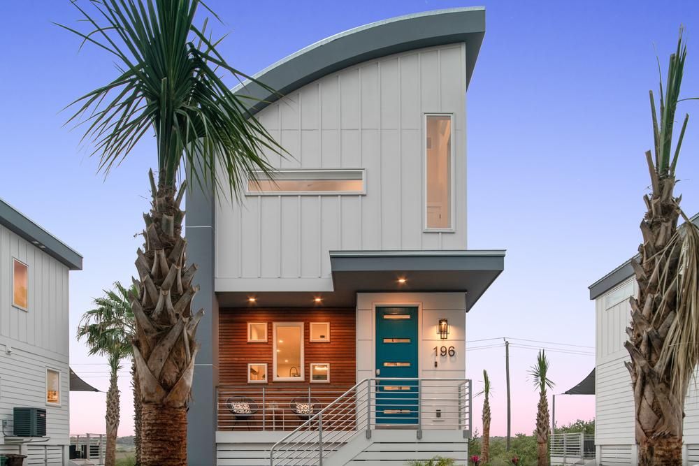 A modern two-story home with a turquoise front door, white siding and an arched gray roof. The front is landscaped with palm trees. 