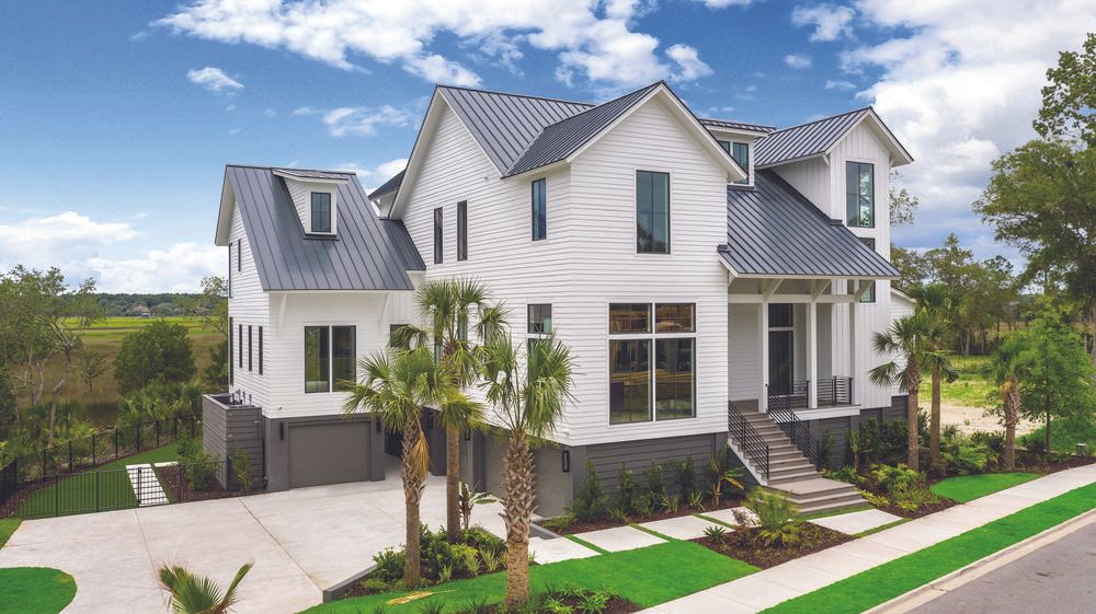 An off-white home decorated in Nichiha Fiber Cement Siding is the focal point. It has dark grey roofing, tall windows, steps leading up to a porch, and a lower-level garage under the home with dark siding. The lawn is full of palm trees, shrubbery, and there is blue sky with white clouds behind it.