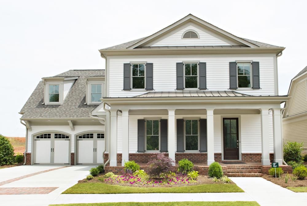 This traditional-style home has white siding, black shutters, a two-car attached garage and a concrete driveway. There is a brick front porch with six pillars. 