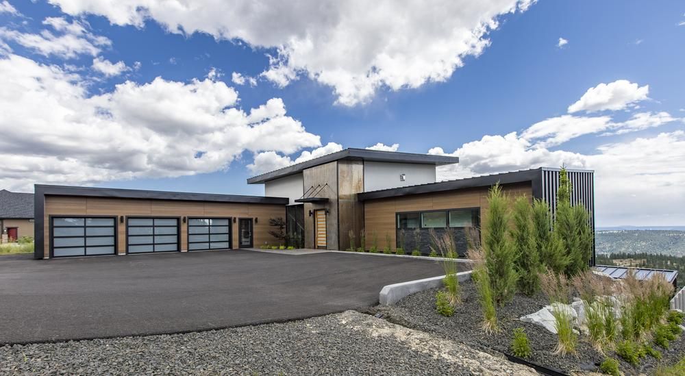 An L-shaped home in front of a large asphalt driveway with fiber cement siding, a black metal roof and a modern design.